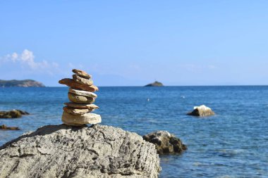 Yunanistan 'ın Skiathos adasındaki bir sahilde büyük ve pürüzlü bir kayanın üzerinde dengeli bir kaya yığını. Parlak, sakin Ege Denizi ve berrak mavi gökyüzünün ardında.