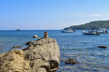 Yunanistan 'ın Kayak Adası' ndaki bir sahilde büyük ve pürüzlü bir kaya yığınının üzerinde dengelenmiş bir kaya yığını. Sakin Ege Denizi 'nde parlak, berrak bir mavi gökyüzü olan teknelerin arkasında..