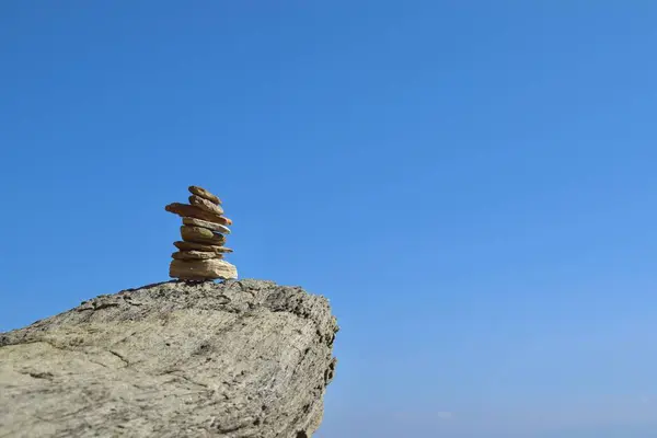 Well-balanced stack of rocks on a large, rough boulder along the Greek coastline, backdropped by a bright, clear blue sky.