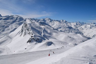 Kayakçılar ve snowboardcular Les Menuires kayak merkezindeki karlı pistlerde yokuş aşağı gidiyorlar. Üç vadideki karlı Fransız dağlarının zirvelerine bakan kış sporları aktiviteleri.