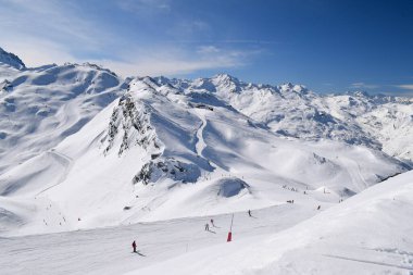 Kayakçılar ve snowboardcular Les Menuires kayak merkezindeki karlı pistlerde yokuş aşağı gidiyorlar. Üç vadideki karlı Fransız dağlarının zirvelerine bakan kış sporları aktiviteleri.