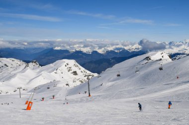 Kayakçılar ve snowboardcular Les Menuires kayak merkezindeki karlı pistlerde yokuş aşağı gidiyorlar. Üç vadideki karlı Fransız dağlarının zirvelerine bakan kış sporları aktiviteleri.
