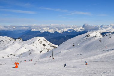 Kayakçılar ve snowboardcular Les Menuires kayak merkezindeki karlı pistlerde yokuş aşağı gidiyorlar. Üç vadideki karlı Fransız dağlarının zirvelerine bakan kış sporları aktiviteleri.