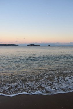 Yunanistan 'ın Skiathos kentindeki kumlu bir plajda gün batımını canlandırın. Gökyüzünde, sakin sahil dalgalarının üzerinde, Yunan ada manzaralarının arka planında, yumuşak pastel renkler parlıyor..