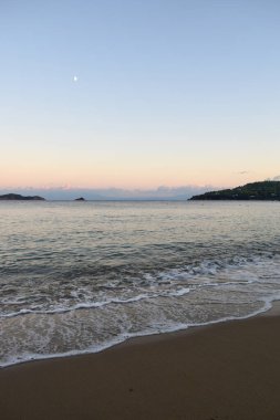 Yunanistan 'ın Skiathos kentindeki kumlu bir plajda gün batımını canlandırın. Gökyüzünde, sakin sahil dalgalarının üzerinde, Yunan ada manzaralarının arka planında, yumuşak pastel renkler parlıyor..