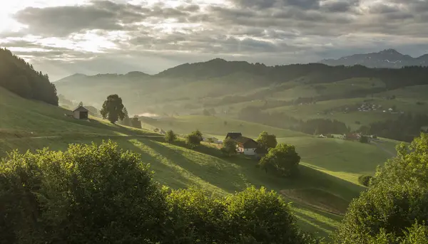 stock image View on the green hills in Appenzeller region. Early morning view with some fog in the valley. Early morning warm morning light. Meadows and some houses on hills in Appenzell, Switzerland.