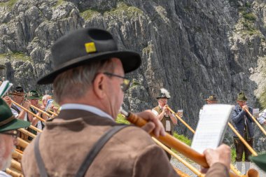 Avusturya 'daki Lnersee dağlık gölünde geleneksel Avusturyalı giysileri içinde erkek ve kadın alphornplayer' lar oynuyor. Arka planda dağ yamacı var. Bir oyuncunun arka planını kapat. 8 Temmuz 2023, Brand, Avusturya