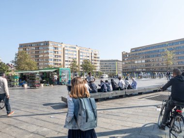 20 Eylül 2024, Elsene, Belçika. Ön planda sırt çantalı insanlar, bisikletli bir adam. Taş bir bankta oturan çok ırklı gençler. Brüksel 'de farklı etnik geçmişleri var. Çiçek ve bitkilerle dolu bir market.