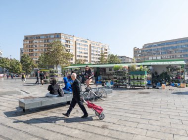 20 september 2024, Elsene, Belgium.  Man walking by with market trolley. Some people sitting on a stone bench. Market stall with flowers and plants in background. Residential buildings. Flagey square. clipart