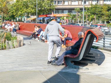 20 Eylül 2024, Elsene, Belçika. Kırmızı ahşap bir bankta oturan farklı etnik kökenli insanlar. Şehir meydanında insanlar ve konut binaları var. Güneşli hava, Brüksel.