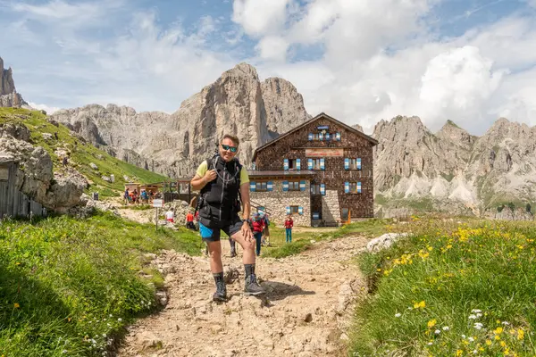 13 Temmuz 2024, Roda di Vael, İtalya. Rotwandhtte 'nin (Rifugio Roda di Val) önünde İtalyan dolomitlerinde dağ kulübesinde yürüyüş yapan beyaz yürüyüşçü. Önde yürüyüş parkuru var. Kulübenin yanında yürüyüşçüler var..