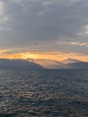 Dalgalanan dalgalarla dolu engin bir denizin üzerinde altın gün batımı. Güneş ışınları dramatik bulutları delip geçer ve uzak dağ zirvelerinde sıcak bir parıltı bırakır. Huzurlu ve manzaralı bir manzara, seyahat, doğa ve rahatlama temaları için mükemmel..