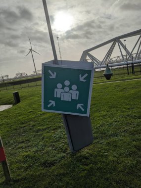 A tilted emergency assembly point sign near the Maeslantkering storm surge barrier in the Port of Rotterdam, Netherlands. The massive floodgate, wind turbine, and cloudy sky contrast with nature, showcasing advanced engineering for coastal protection clipart