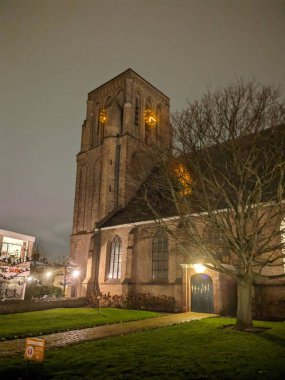 A historic church in the Netherlands illuminated at night. The tall brick tower stands against the dark sky, with glowing lights shining through its arched windows. A peaceful scene with streetlights casting a warm glow on the surrounding greenery. clipart