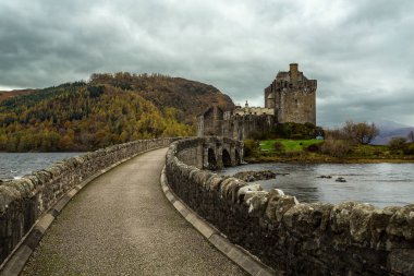 Dornie yakınlarında bulunan eski İskoç şatosu Eilean Donan, kaleye giden taş bir köprü ve altından bir nehir geçiyor. Yamacında ağaçlar olan bir tepe ve arka planda bulutlu bir gökyüzü..