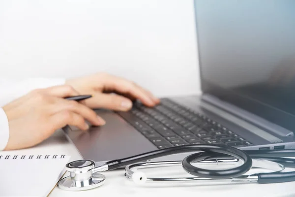 Doktorun çalışma yerinde dizüstü bilgisayar, not defteri ve stetoskop var. Dizüstü bilgisayar kullanan kadın doktor elleri. Teletıp kavramı
