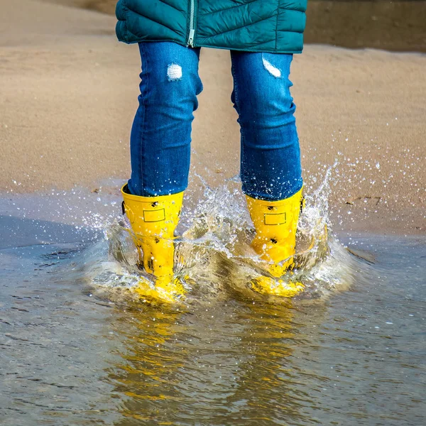 girl in denim jeans with yellow rubber wellington boots splashing  in a puddle on the beach.