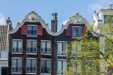 Geleneksel, tarihi Amsterdam şehir evleri, kanalların kıyısında, kantilleri ve kancaları olan, ilkbaharda dar eğimli, güzel mavi gökyüzü olan evlerin ön köşelerinde.