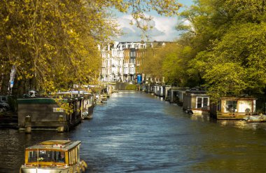 İlkbaharda Amsterdam. Amsterdam Kanalı 'nda su tekneleri var. Her iki tarafında ağaçlar ve parlak gökyüzü.