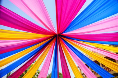Parlak renkli mavi, sarı ve pembe kumaş iplikleri dekoratif bir açık hava örtüsü oluşturarak neşeli ve neşeli bir atmosfer yaratıyor, güneşli ortamlarda etkinlikler ve kutlamalar için ideal..