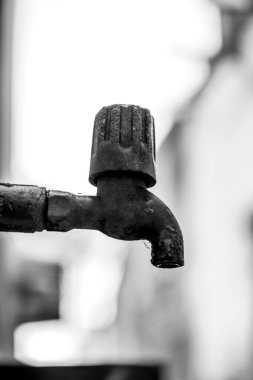 Eski bir açık hava metal su musluğunun siyah beyaz fotoğrafı, kırsal dokusunu ve zamansız tasarımını bulanık bir arka plana karşı sergiliyor, endüstriyel çürümeyi ve minimalist estetiği vurguluyor..