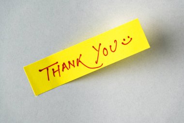 El yazısıyla yazılmış bir teşekkür mesajı ve yumuşak, tarafsız bir yüzeye yazılmış gülen bir yüzle parlak sarı bir not. Minnettarlığı, pozitifliği ve kişisel ifadeyi temsil eder.