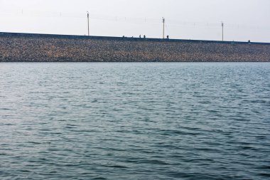 İnce dalgalar ve sakin bir ortam ile vurgulanmış, doğanın ve insan mühendisliğinin bir karışımını yakalayan, çakıl barajına yakın büyük bir su deposunun panoramik görüntüsü..