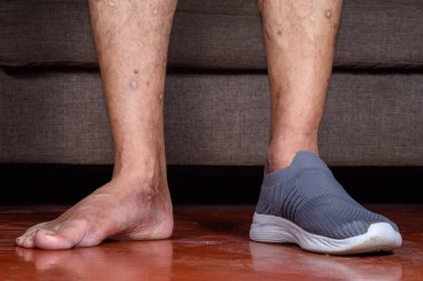 Birinin bacakları yakın plan, biri gri ayakkabılı, diğeri çıplak, ahşap zeminde duruyor, ayakkabı ve deri detaylarını sıradan bir ortamda sergiliyor..