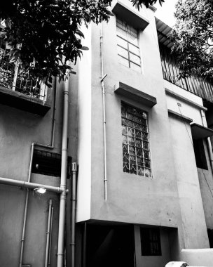 Büyük pencereler, yapısal borular ve ağaç dalları tarafından çerçevelenmiş mimari elementlerin yer aldığı bir binanın tek renkli fotoğrafı.