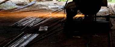 Endüstri sahnesinde, kırsal bir atölyede metali kıvılcımlarla kesen bir işçi görülüyor. Görüntü, maddelerin şekillendirilmesinde kullanılan el işçiliğini vurgular ve metal, doğa ve zanaatkarlığın zıt unsurlarını vurgular..