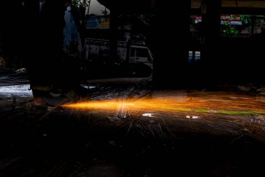 Endüstri sahnesinde, kırsal bir atölyede metali kıvılcımlarla kesen bir işçi görülüyor. Görüntü, maddelerin şekillendirilmesinde kullanılan el işçiliğini vurgular ve metal, doğa ve zanaatkarlığın zıt unsurlarını vurgular..