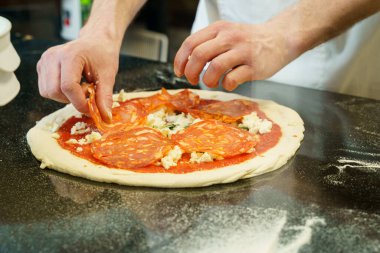 Bir şef domates soslu ve mozzarella peynirli pizza hazırlıyor, pizzanın üzerine salam yayıyor.