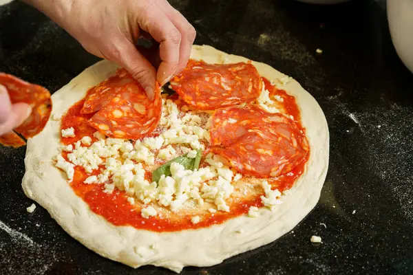 Bir şef domates soslu ve mozzarella peynirli pizza hazırlıyor, pizzanın üzerine salam yayıyor.