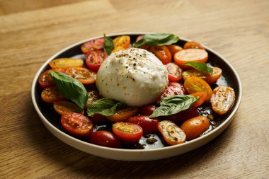 Bir tabak taze burrata peyniri. Etrafı canlı domates yarısıyla çevrili ve fesleğen yapraklarıyla süslenmiş. Basit ama renkli sunum malzemelerin tazeliğini vurgular, hafif ve lezzetli yemekler için mükemmeldir..