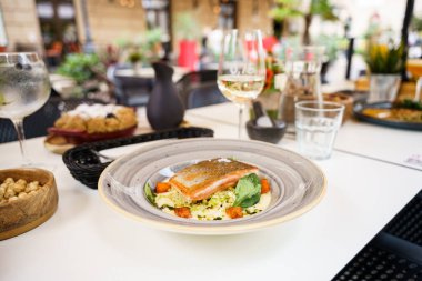 A beautifully plated grilled salmon dish served with a mix of vegetables and sauce in an outdoor restaurant setting. The scene includes various drink glasses and a decorative tableware. clipart