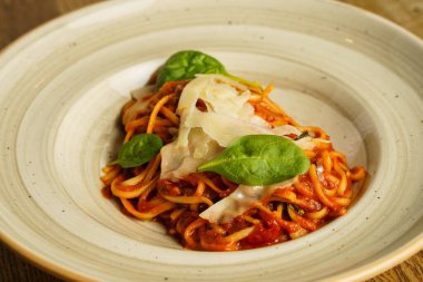 Taze fesleğen yaprakları ve tıraş edilmiş parmesan peyniri ile süslenmiş domates soslu güzel bir tabak spagetti. Yemek kırsal bir seramik kasede sunuluyor, sıcak ve davetkar bir sunum yaratıyor..
