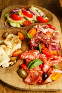 Pişmiş et, peynir, domates ve salatalıklı taze salata ve bir sepet dilimlenmiş ekmek içeren kırsal bir şarküteri tahtası. Yayılım, hafif bir aperatif ya da aperatif olarak paylaşmak için ideal doku ve tatlar sunuyor..