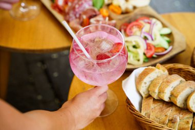 Buzlu ve biberiyeli ferahlatıcı bir kokteyl bardağı tutan bir el. Arka planda, ahşap bir masaya yerleştirilmiş et, peynir ve sebzelerle dolu bir şarküteri tahtası var..