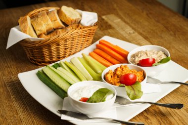 Çeşitli soslar ve bir sepet dilimlenmiş ekmeğin yanında havuç, kereviz ve salatalık çubuklarıyla servis edilen taze bir sebze tabağı. Bu sağlıklı aperatif seti kırsal bir ahşap masada sergileniyor..