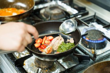 Bir şef içinde ton balığı, kiraz domatesleri ve pesto sosu olan bir tavaya sıvı ekler. Canlı malzemeler bir gaz sobasında kaynıyor ve lezzetli bir yemek hazırlama yolunda bir adım atıyor..