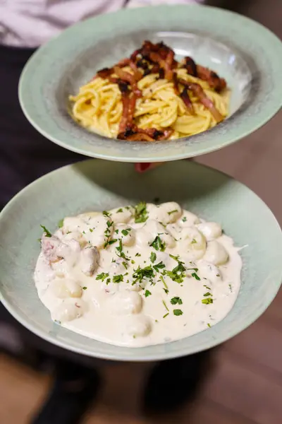 Zarif pastel yeşil kaselerde servis edilen iki tabak spagetti carbonara üstü çıtır pancetta ve maydanozla süslenmiş kremalı gnocchi..