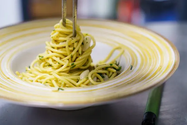 Profesyonel bir mutfakta maşa kullanan bir şefin spagetti aglio ve olio tabaklarını dikkatlice kaplarken çekilmiş yakın plan bir fotoğrafı. Makarna zeytinyağı, sarımsak ve taze maydanozla kaplıdır..