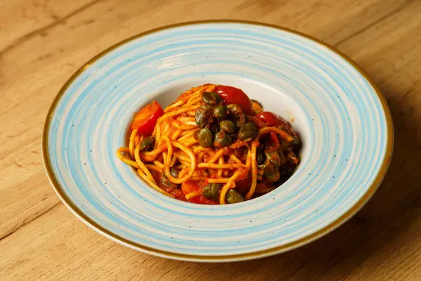 Zengin domates soslu bir tabak spagetti, kiraz domatesleri ve kapari ahşap bir masanın üzerinde mavi çizgili seramik bir tabakta zarif bir şekilde servis edilir. Yemek Akdeniz lezzetlerine vurgu yapıyor.
