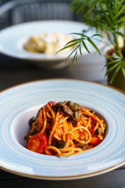 Zengin domates soslu bir tabak spagetti, kiraz domatesleri ve kapari ahşap bir masanın üzerinde mavi çizgili seramik bir tabakta zarif bir şekilde servis edilir. Yemek Akdeniz lezzetlerine vurgu yapıyor.