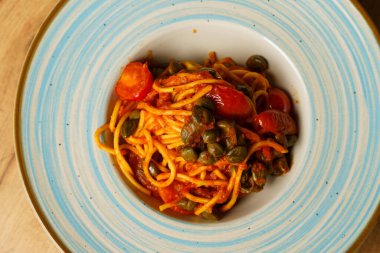 Zengin domates soslu bir tabak spagetti, kiraz domatesleri ve kapari ahşap bir masanın üzerinde mavi çizgili seramik bir tabakta zarif bir şekilde servis edilir. Yemek Akdeniz lezzetlerine vurgu yapıyor.