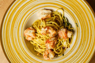 Ahşap bir masanın üzerinde sarı çizgili seramik bir tabakta güzel bir kaplanmış karides spagetti aglio e olio sunulur. Makarna zeytinyağı, sarımsak ve taze bitkilerle kaplıdır..