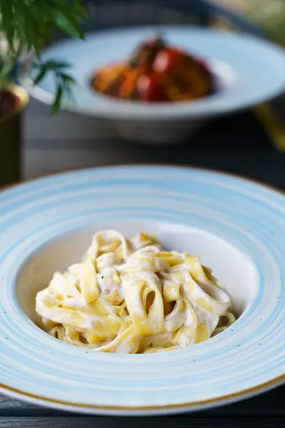 Mavi çizgili seramik tabakta servis edilen zengin ve kremsi peynir soslu fettuccine alfredo makarnası. Bulanık bir arka planda başka bir kaplama ve yeşil bitkiler.
