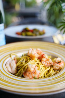 Hafif sarımsaklı ve zeytinyağı soslu yumuşak karidesli ve taze otlarla süslenmiş bir tabak linguine makarna. Tabak, koyu ahşap bir masada seramik bir kasenin içinde..