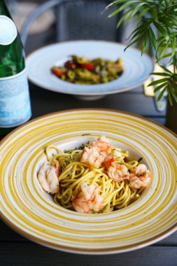 Hafif sarımsaklı ve zeytinyağı soslu yumuşak karidesli ve taze otlarla süslenmiş bir tabak linguine makarna. Tabak, koyu ahşap bir masada seramik bir kasenin içinde..