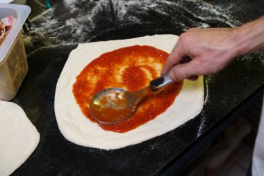 Şefin elinin, kepçeyle çiğ pizza hamuruna taze domates sosu sürerken yakın plan görüntüsü. Profesyonel bir mutfakta geleneksel pizza yapma sürecini vurgulamak